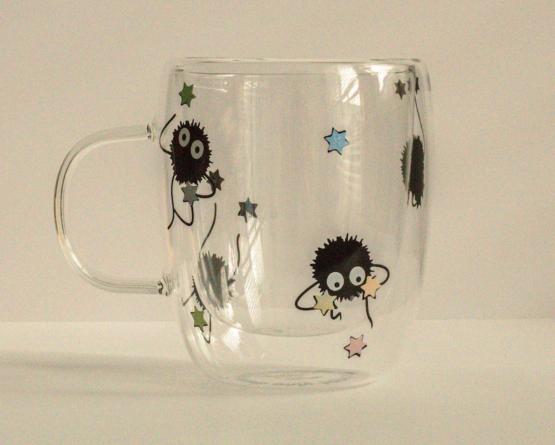 May include: A clear glass mug with a double wall design features black and white illustrations of fuzzy creatures with large eyes and stars. The mug has a handle.