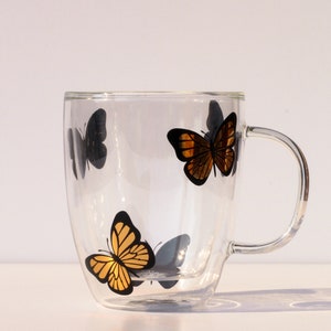 May include: A double-walled glass mug with a handle features a gold butterfly design. The mug has a clear glass exterior and a black butterfly design on the inside.