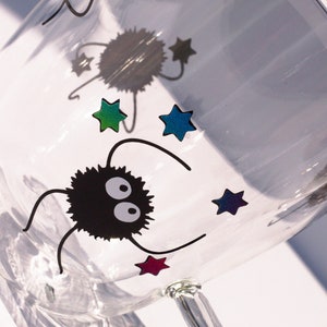 May include: A clear glass mug with a black and white cartoon character and colorful stars. The character has two large eyes and is fuzzy.