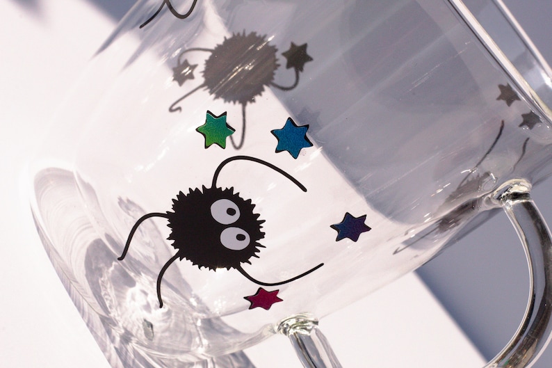 May include: A clear glass mug with a black and white cartoon character and colorful stars. The character has two large eyes and is fuzzy.