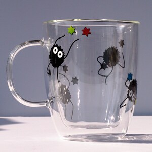 May include: A clear glass double-walled mug with a black handle. The mug has a design of black cartoon characters with white eyes and stars in various colors.