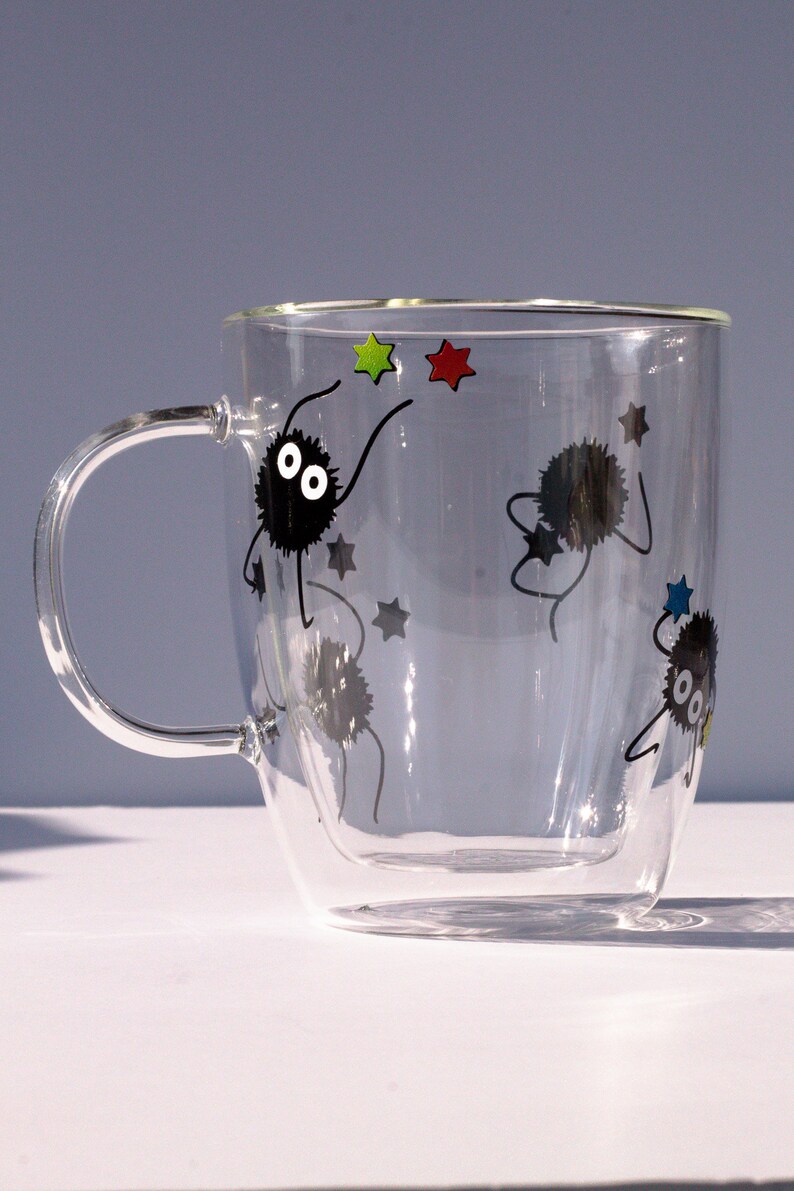 May include: A clear glass double-walled mug with a black handle. The mug has a design of black cartoon characters with white eyes and stars in various colors.