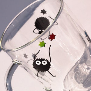 May include: A clear glass mug with a black handle features a design of two black fuzzy creatures with large eyes and red, green, and white stars.