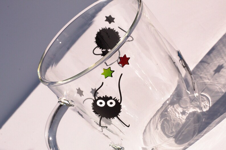 May include: A clear glass mug with a black handle features a design of two black fuzzy creatures with large eyes and red, green, and white stars.