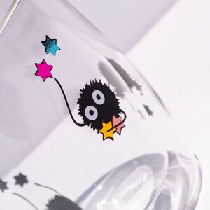 May include: A clear glass with a black and white cartoon character with two large eyes. The character is holding a pink and yellow star. There are other stars in the background, including a pink, blue, and yellow star.