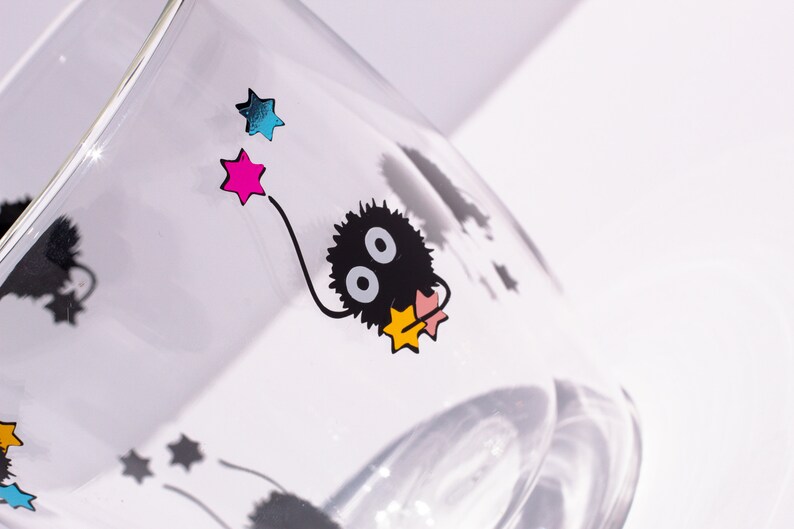 May include: A clear glass with a black and white cartoon character with two large eyes. The character is holding a pink and yellow star. There are other stars in the background, including a pink, blue, and yellow star.