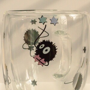 May include: A clear glass mug with a black and white cartoon character design. The character is a fluffy, round creature with large eyes and a pink bow. The mug has a handle and is decorated with stars.