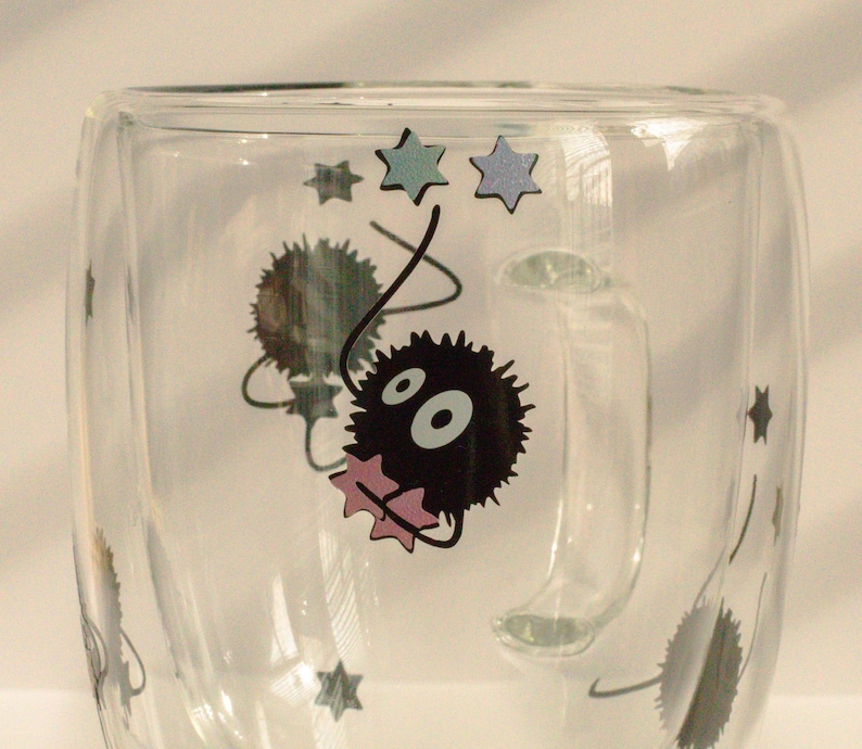 May include: A clear glass mug with a black and white cartoon character design. The character is a fluffy, round creature with large eyes and a pink bow. The mug has a handle and is decorated with stars.