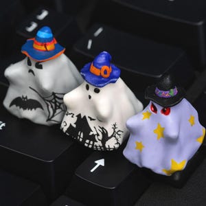May include: Three Halloween ghost figurines, each with a different design and hat. One ghost has a bat and web, another a haunted house, and the last yellow stars. The ghosts are white, with hats in blue, orange, and black.