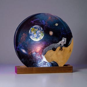 May include: A unique resin art piece depicting an astronaut gazing at Earth within a galaxy scene. The artwork features a wooden base and a semi-circular design with a vibrant display of planets and stars. The astronaut is resting on a rock formation.