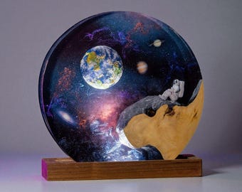 Könnte beinhalten: Ein einzigartiges Kunstwerk aus Harz, das einen Astronauten zeigt, der die Erde in einer Galaxieszene betrachtet. Das Kunstwerk hat einen Holzsockel und ein halbkreisförmiges Design mit einer lebendigen Darstellung von Planeten und Sternen. Der Astronaut ruht auf einer Felsformation.