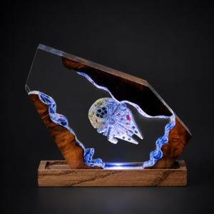 May include: A unique, illuminated sculpture featuring a Millennium Falcon model encased in clear resin. The resin is set within a wooden frame with a blue light accent. The base is made of wood.