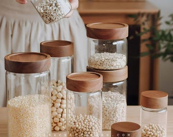 May include: A collection of clear glass storage jars with cylindrical wooden lids, filled with various grains and legumes. The jars are arranged in different sizes, and one jar is held up, displaying its contents clearly.
