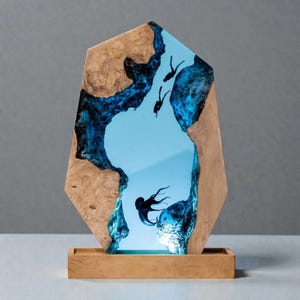 May include: A unique wooden art piece featuring a blue resin river scene with silhouettes of divers and an octopus. The artwork is set in a wooden frame and sits on a wooden base. The piece is approximately 12 inches tall.
