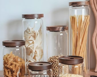 May include: A set of six clear glass storage canisters with dark brown wooden lids. The containers hold various dry food items including rotini pasta, flat pasta shapes, soybeans, small white beans, and grains, arranged in different heights.