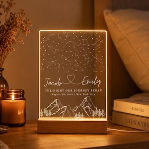 May include: A rectangular acrylic night light with a warm LED glow, featuring an engraved star map, a heart icon, the names Jacob and Emily, the text THE NIGHT OUR JOURNEY BEGAN, August 08, 2025, New York City, and a mountain and forest landscape.