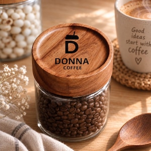 Op de afbeelding: Een glazen voorraadpot gevuld met donker gebrande koffiebonen, voorzien van een rond houten deksel gegraveerd met een koffiekop-icoon, de naam DONNA en het woord COFFEE. Het deksel heeft een natuurlijke houtnerf en een bruine afwerking.