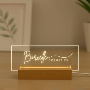 May include: A lighted sign with the text "Borcelle COSMETICS" in a script font. The sign is rectangular and made of clear acrylic, resting on a wooden base. The sign is illuminated with a warm, soft glow.