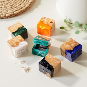 May include: A collection of small, square ring boxes with wooden lids and colorful resin bases. The boxes feature shades of orange, blue, green, white, and black. One box is open, revealing a ring inside. The boxes are displayed on a white surface.