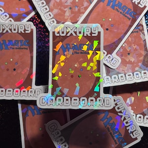 May include: A close-up of a holographic sticker with the text "LUXURY" and "CARDBOARD" printed on it. The sticker features a Magic: The Gathering logo and a design of holographic confetti.