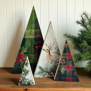 May include: A collection of decorative wooden Christmas trees in various sizes. The trees feature festive designs, including plaid patterns, a deer illustration, and botanical elements with red berries and pine needles. The trees are arranged on a wooden surface.