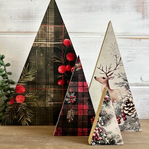 May include: Four triangular wooden decorations of varying heights. The largest features a dark green plaid pattern with red berries and pine needles. The medium piece has a red and black plaid design. The others feature a deer, pinecones, and berries.