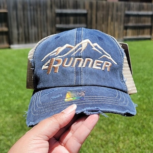May include: A blue and brown distressed trucker hat with a brown embroidered mountain range and the text "4RUNNER".