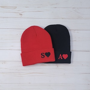 May include: Two knit beanies, one red and one black, with a heart and letter embroidered on each. The red beanie has a black "S" and a black heart, and the black beanie has a white "A" and a red heart.