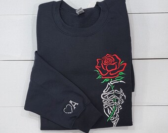 May include: Black sweatshirt with a white skeleton hand holding a red rose with green leaves. The left sleeve has a white heart with the letter "A" inside.