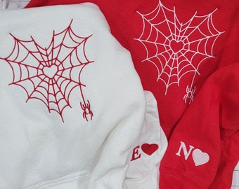 May include: A red hooded sweatshirt and a white hooded sweatshirt, each featuring an embroidered heart-shaped spider web design with a small spider. The sleeves are embroidered with the letters E and N, each accompanied by a small heart icon.