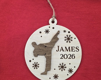 May include: A round, light-colored wooden ornament featuring a dark wood cutout of a person performing a martial arts kick. The ornament is engraved with the name JAMES and the year 2026, surrounded by snowflake cutouts and a jute twine hanger.