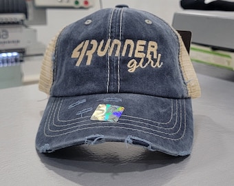 May include: A distressed navy blue baseball cap with tan mesh side panels. The front features embroidered text that reads 4RUNNER girl in a light tan thread. The brim has frayed edges and a small holographic sticker centered on the front panel.