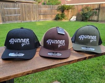May include: Three trucker hats with a black mesh back and a front panel in black, brown, and green. Each hat has a white embroidered logo that says "4Runner girl".
