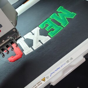 May include: A close-up of an embroidery machine stitching the word "MEXIC" in green, white, and red thread onto a black Gildan t-shirt. The machine is labeled "13 x 16" Mighty Hoop".