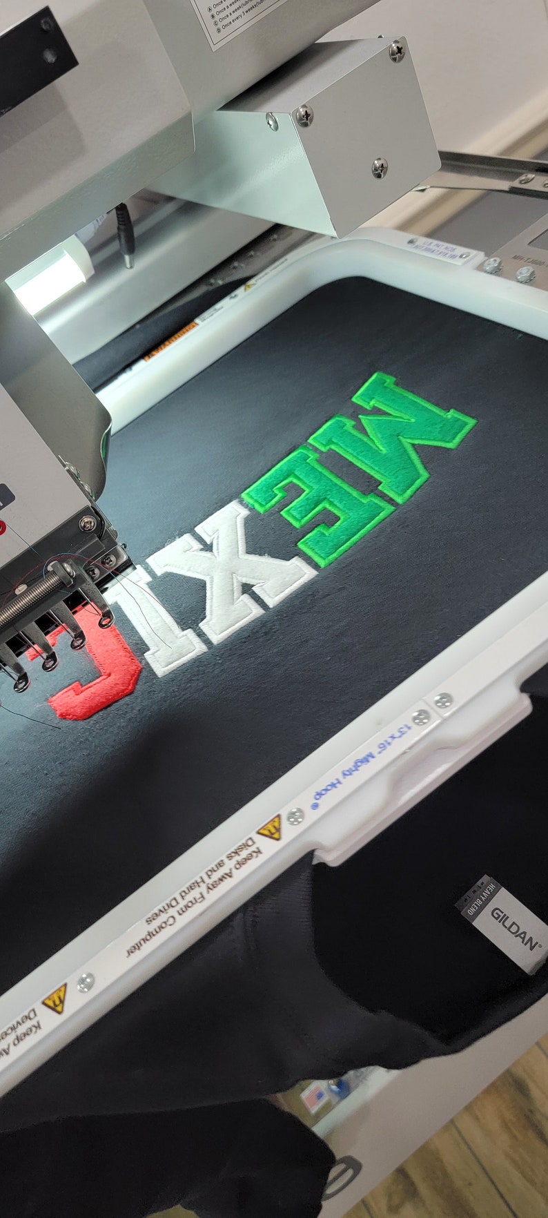 May include: A close-up of an embroidery machine stitching the word "MEXIC" in green, white, and red thread onto a black Gildan t-shirt. The machine is labeled "13 x 16" Mighty Hoop".