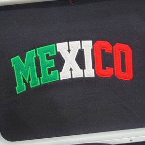 May include: A black fabric with the word "MEXICO" embroidered in green, white, and red letters. The fabric is in a 13x16 inch embroidery hoop.