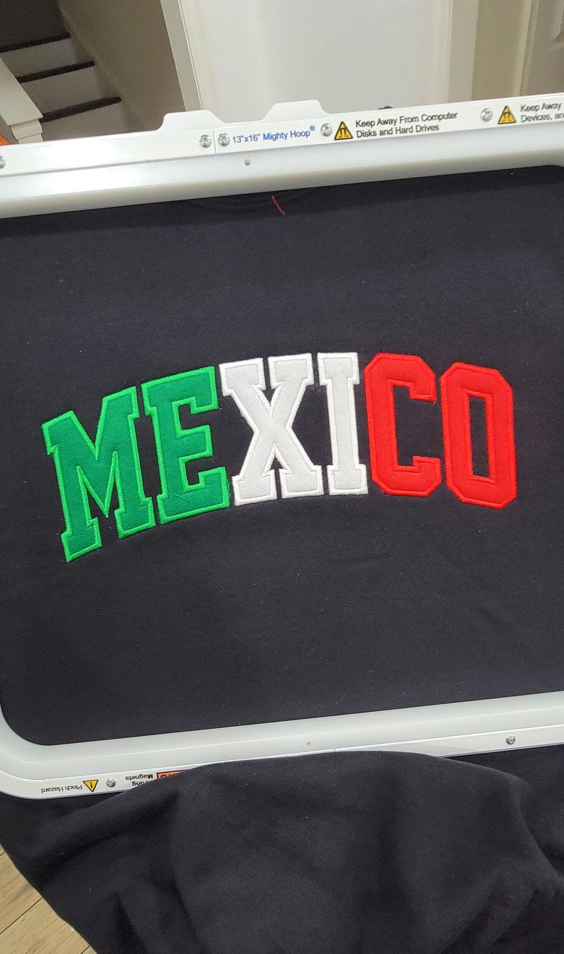 May include: A black fabric with the word "MEXICO" embroidered in green, white, and red letters. The fabric is in a 13x16 inch embroidery hoop.