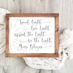 Pode incluir: Um sinal enquadrado em madeira com fundo branco e texto preto que diz "Travel light, live light, spread the light, be the light. - Yogi Bhajan". O sinal tem um design abstrato bege no fundo.