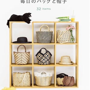 以下が含まれることがあります： 「エコアンダリヤで編む 毎日のバッグと帽子 32 items」というタイトルの手芸本。9つのマスがある明るい木製の棚に、様々な編み込みバッグ、帽子、黒い猫のぬいぐるみ、ピンクの花が入ったガラスの花瓶、オレンジジュースが入ったグラスが並んでいる。