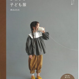 Puede incluir: Una portada de libro con el título "Osharete Kawaii Kodomo Fuku" en japonés, que se traduce como "Ropa de niños elegante y linda". La portada presenta a una niña pequeña con una blusa gris con cuello blanco y pantalones marrones. Ella está sosteniendo una taza de papel en su oído, como si estuviera hablando por teléfono. El libro es parte de la "Heart Warming Life Series" y es publicado por "Nihon Vogue-sha".