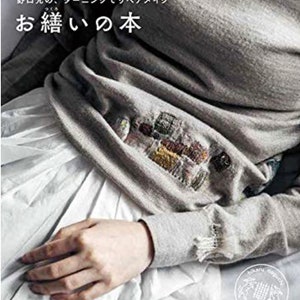 Può includere: Un libro intitolato "Otsukuri no Hon" (Il libro della cucitura) con un primo piano di una mano su un maglione grigio con ricami colorati. Il libro tratta della riparazione dei vestiti usando tecniche di rammendo.