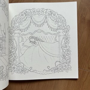 Op de afbeelding: Een zwart-wit kleurboekpagina met een lijntekening van een persoon die in een bed slaapt onder een rozenbaldakijn. De sc&egrave;ne wordt omlijst door een boog van bloeiende rozen en bladeren, met aan de linkerkant een kasteel zichtbaar.