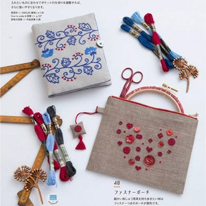 May include: A linen sewing case with blue floral embroidery and a button closure, next to a zippered pouch with a heart design made of red buttons and embroidery. Also shown are embroidery floss, scissors, and a wooden embroidery hoop.