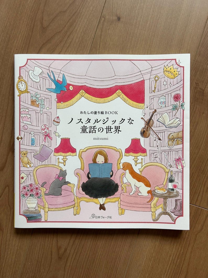 Op de afbeelding: Een kleurboek getiteld "わたしの塗り絵BOOK ノスタルジックな童話の世界" door mitsumi, met een centrale illustratie van een persoon die in een stoel leest, omringd door roze fauteuils, een kat, een hond, lampen en planken vol met diverse voorwerpen.