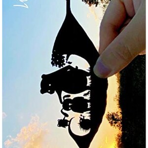 May include: A black paper silhouette of a leaf with a family of bears inside. The silhouette is held up against a sunset sky.