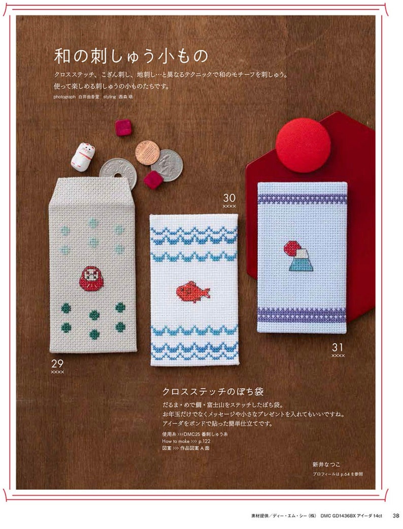 May include: A page from a Japanese craft book featuring cross-stitch designs. The image showcases three small, embroidered pouches with different motifs: a Daruma doll, a red fish, and Mount Fuji. The text includes Japanese characters and instructions.