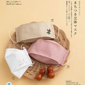 May include: A page from a craft book featuring three handmade face masks. The masks are beige, pink, and white, each with embroidered designs. The beige mask has a sprig of red berries, the pink mask has a floral design, and the white mask has a geometric pattern.