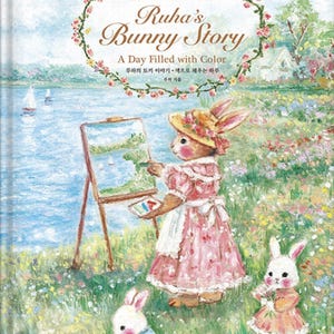 May include: A hardback book titled Ruha's Bunny Story: A Day Filled with Colour, featuring an oil pastel illustration of three rabbits in a field by a lake. One rabbit paints on an easel, while two others sit in the grass. The cover includes floral accents.