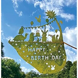 May include: A green leaf with a happy birthday message cut out of it. The leaf is decorated with stars and a bunny rabbit. The leaf is held up against a blue sky with white clouds.
