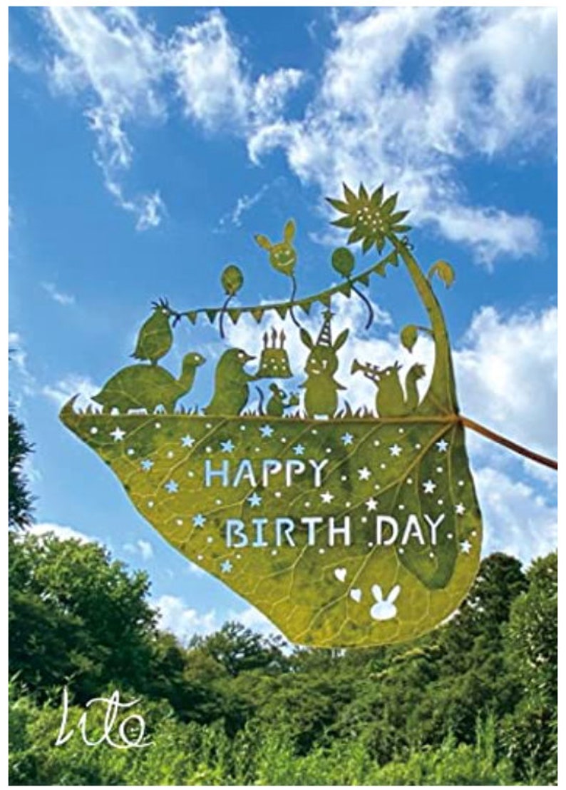 May include: A green leaf with a happy birthday message cut out of it. The leaf is decorated with stars and a bunny rabbit. The leaf is held up against a blue sky with white clouds.